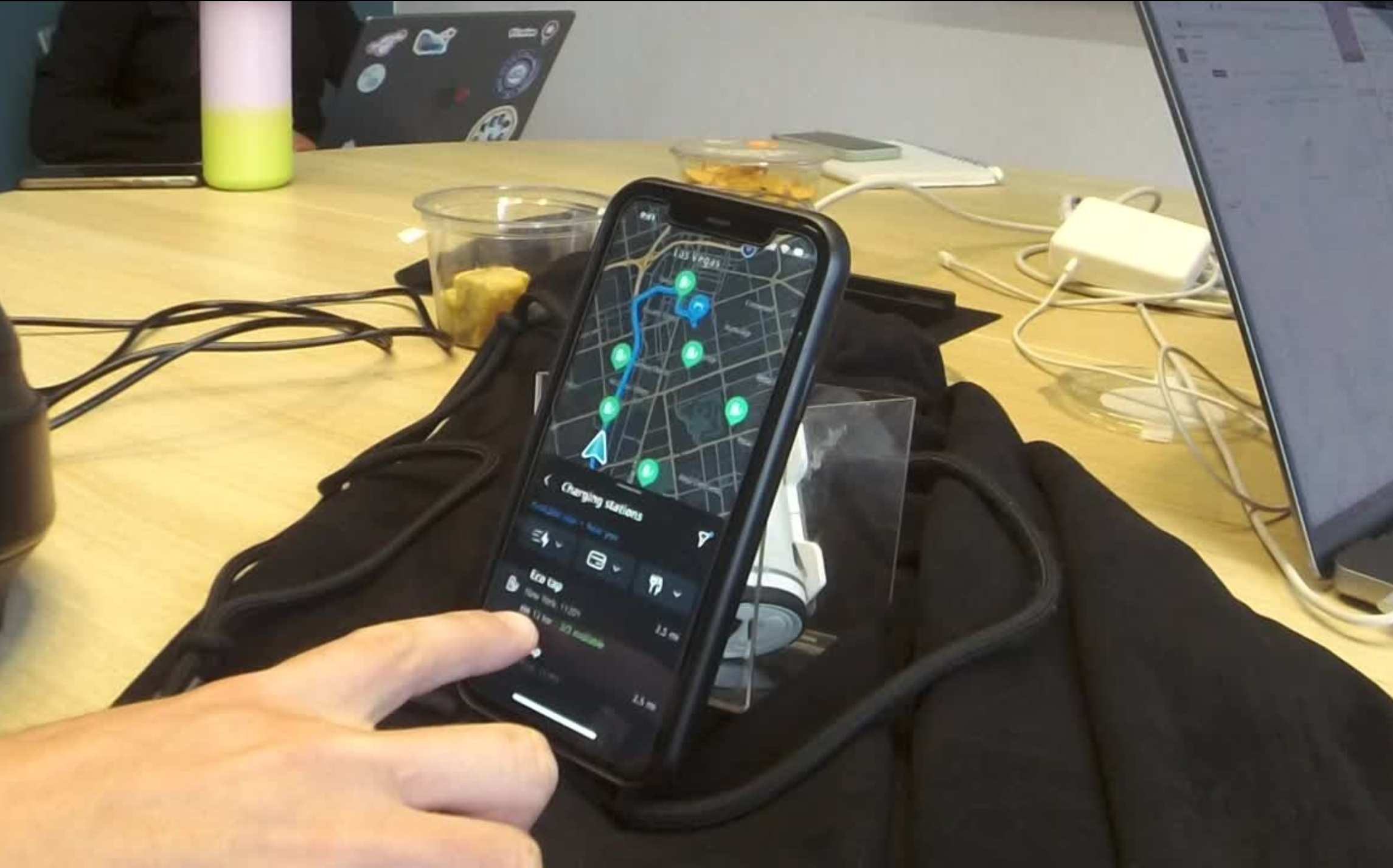 Usability test participant holding a phone showing the charging station search results while seated at a desk with a simulated driving setup